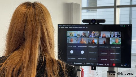 Eine rothaarige Frau sitzt mit dem Rücken zur Kamera und guckt auf einen Bildschirm. Auf dem Bildschirm ist eine Videokonferenz zu sehen. 