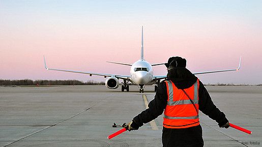 Ein Fluglotse in einer roten Warnweste steht mit dem Rücken zur Kamera. Weit vor ihm ist ein Flugzeug zu sehen, das auf der Flugbahn steht.