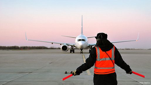 Ein Fluglotse in einer roten Warnweste steht mit dem Rücken zur Kamera. Weit vor ihm ist ein Flugzeug zu sehen, das auf der Flugbahn steht.
